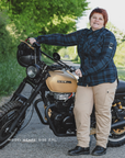 a woman standing next to a Honda motorcycle on a road with greenery in the background wearing blue lumberjack motorcycle shirt from Shima