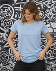 Woman wearing a light blue t-shirt with text against a graffiti-covered wall.