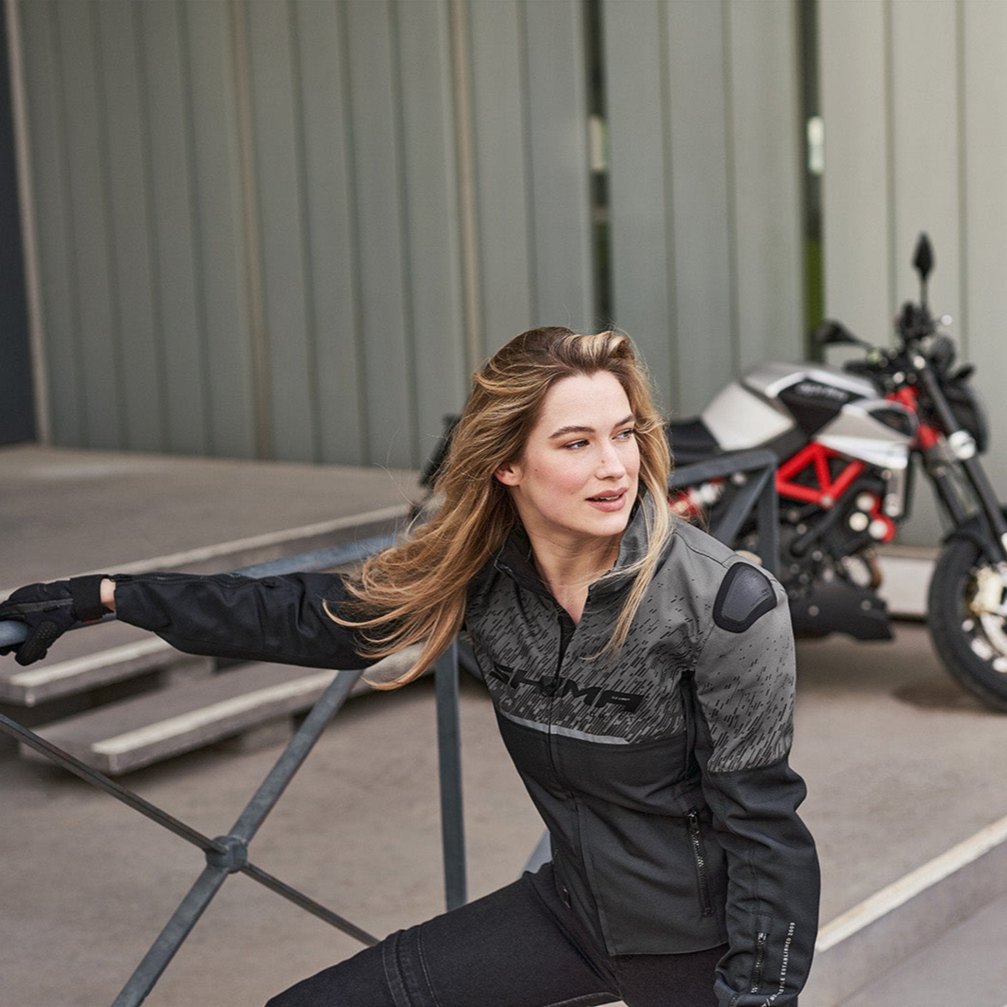 A blond woman near the motorcycle wearing grey women's motorcycle jacket from SHIMA