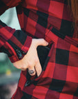 Close-up of a woman wearing a red and black flannel-style women's jacket, putting a phone in the side pocket.