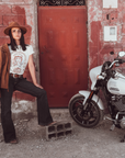 Woman standing next to a Motorcycle wearing a white T-shirt with a Woman wearing a Motorcyclehelmet print and the text "Wild dust sister" and "Enjoy The ride" in orange