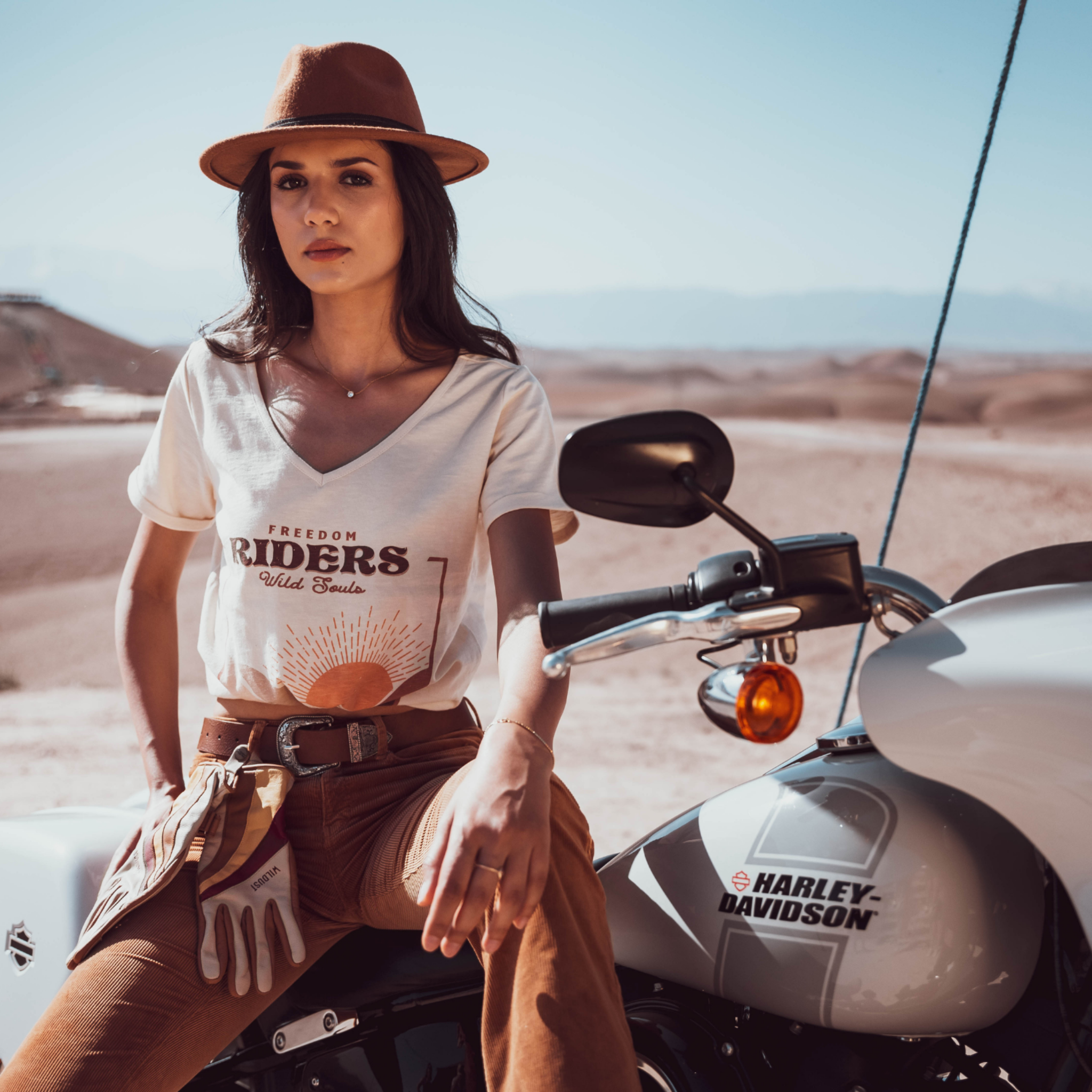 Woman sitting on a Motorcycle wearing a T-shirt with a sunset/sunrise print and the text &quot;Freedom Riders Wild Souls&quot; and &quot;Wildust sisters&quot;