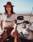 Woman sitting on a Motorcycle wearing a T-shirt with a sunset/sunrise print and the text "Freedom Riders Wild Souls" and "Wildust sisters"