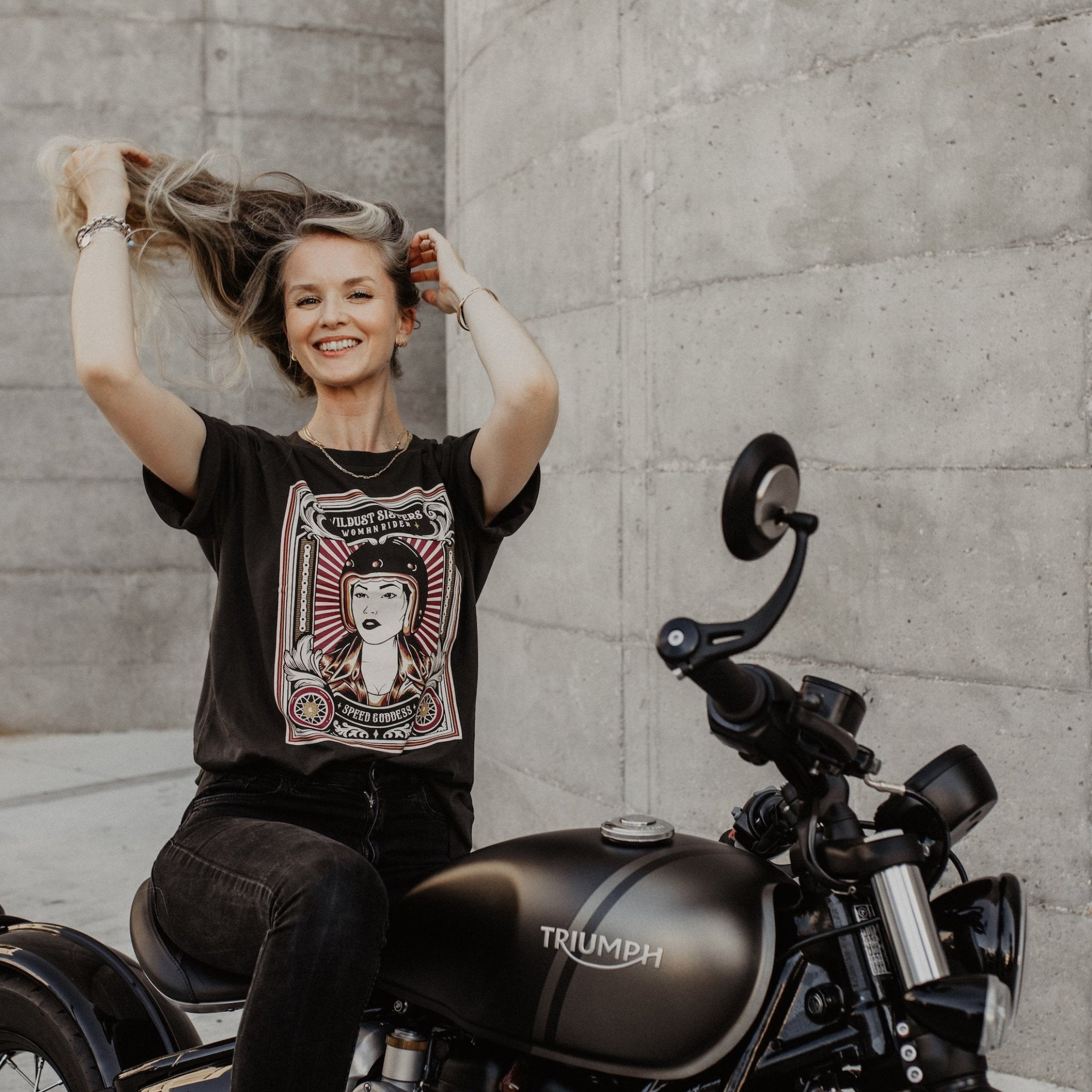 Woman sitting on a Motorcycle wearing  a T-shirt with a women wearing a Motorcyclehelmet print and with the text &quot;Wild dust sisters woman rider&quot; and &quot;Speed Goddess&quot;