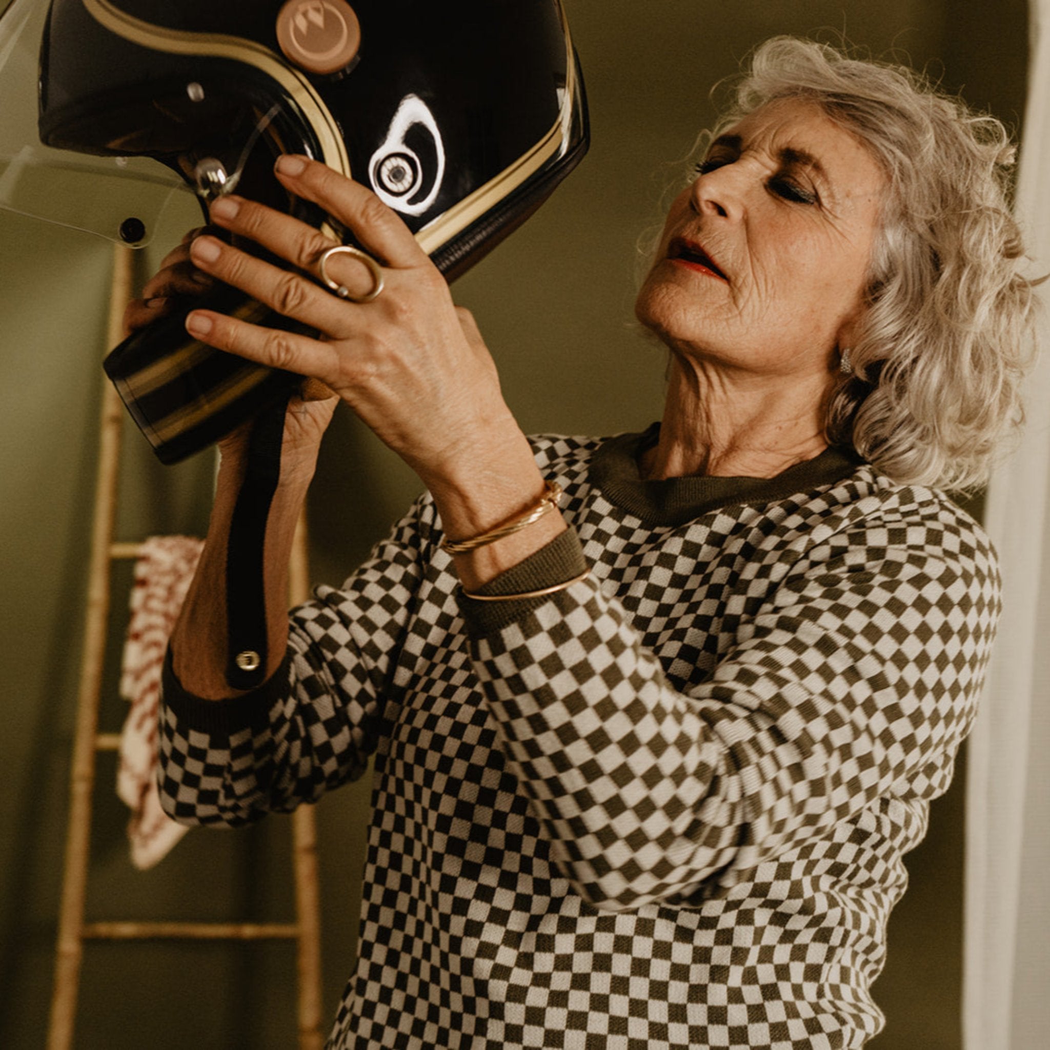 an older lady wearing a knitted khaki green and white jumper with chessboard motives from Wildust putting on her helmet