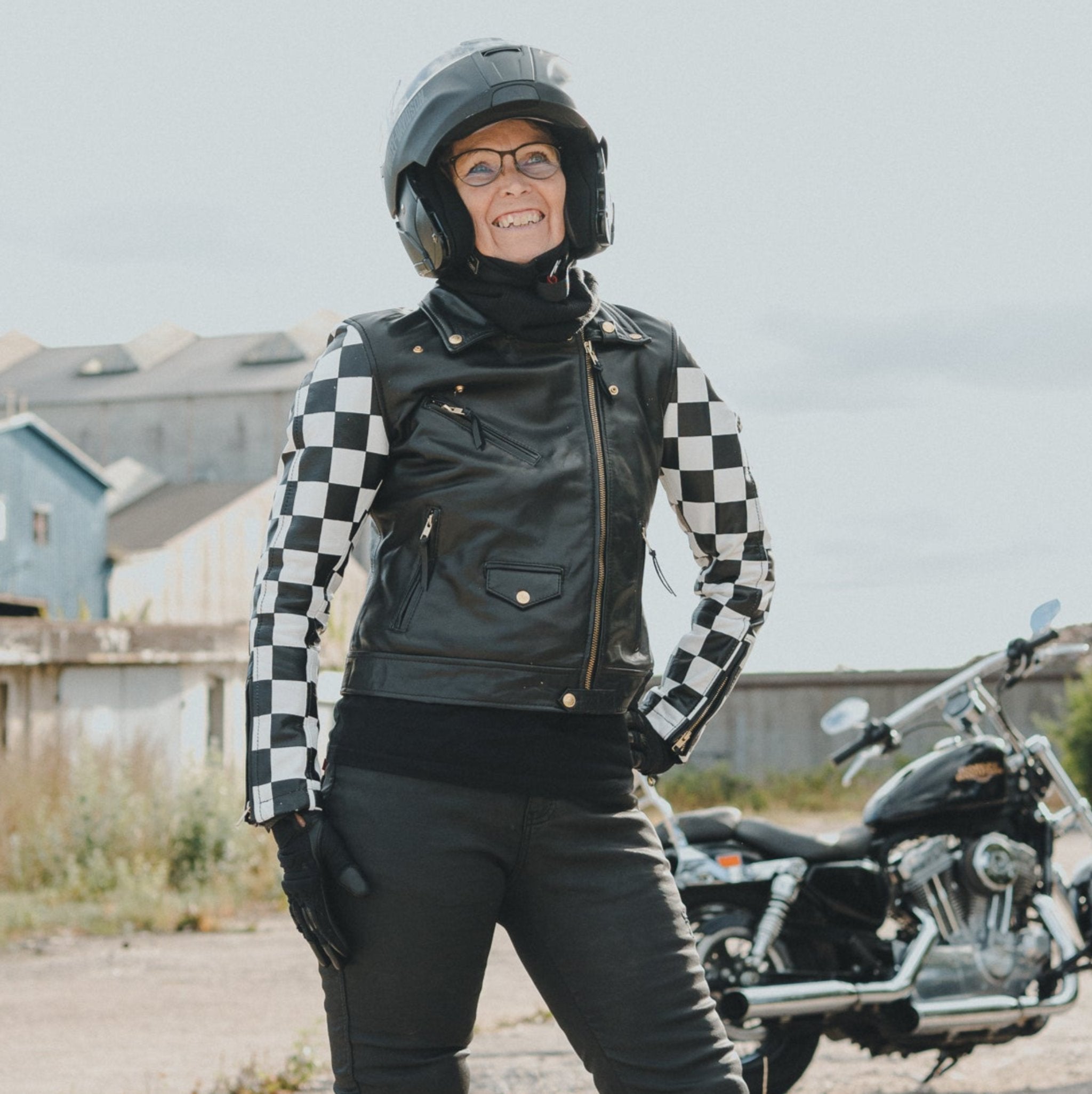 Smiling woman standing outside in front of a motorcycle, wearing motorcycle clothing and a black knitted neck warmer from Moto Girl.