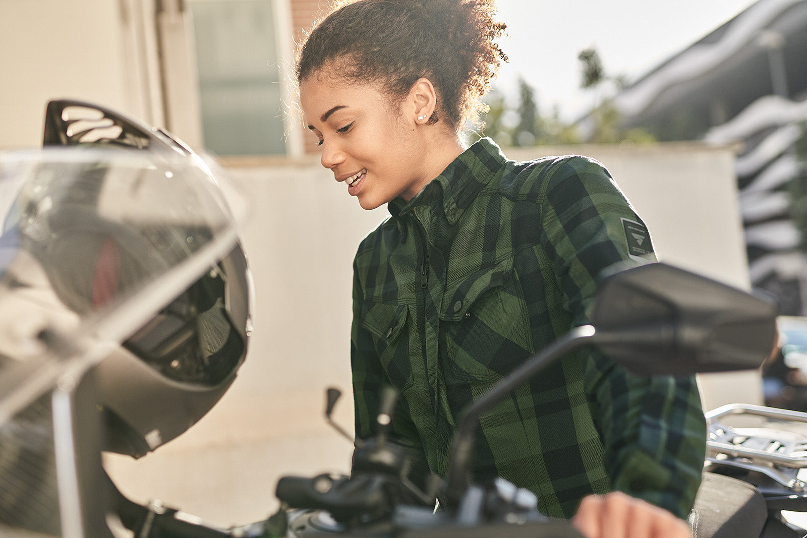 Woman sitting on a motorcycle, wearing a green plaid shirt, with a blurred background.
