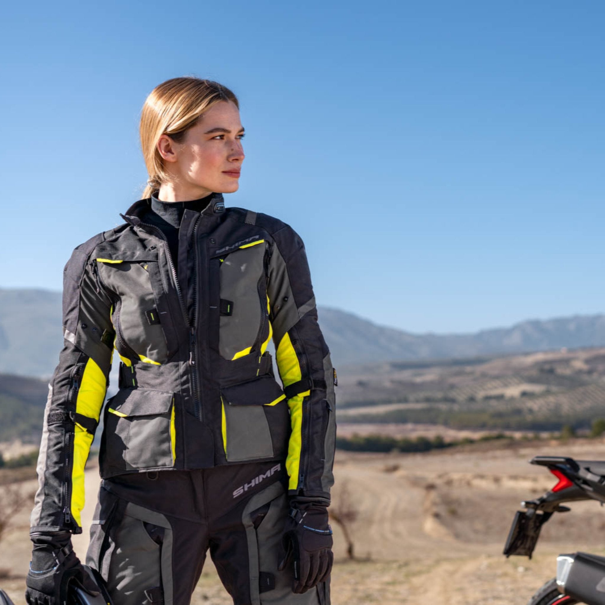 A woman standing by her mc wearing shima motorcycle touring jacket for women in black/ fluo