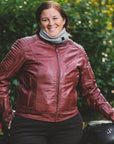 Woman in a red Valerie jacket and light gray neck warmer standing outside next to a Suzuki motorcycle.