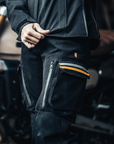 Person wearing black motorcycle pants with zippers and reflective stripes, standing next to a motorcycle.