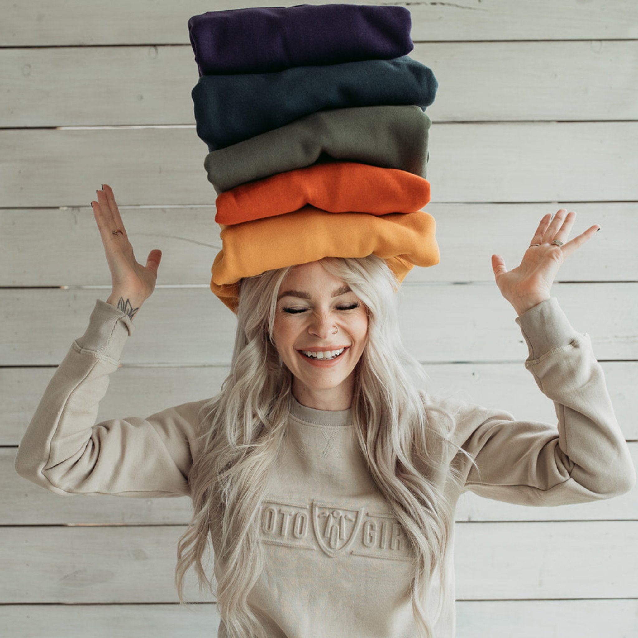 A young lady wearing sand colour motorcycle sweatshirt with Moto Girl logo and holding 5 sweatshirts on her head