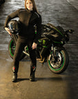 A young woman standing by the motorcycle wearing black motorcycle protective clothes