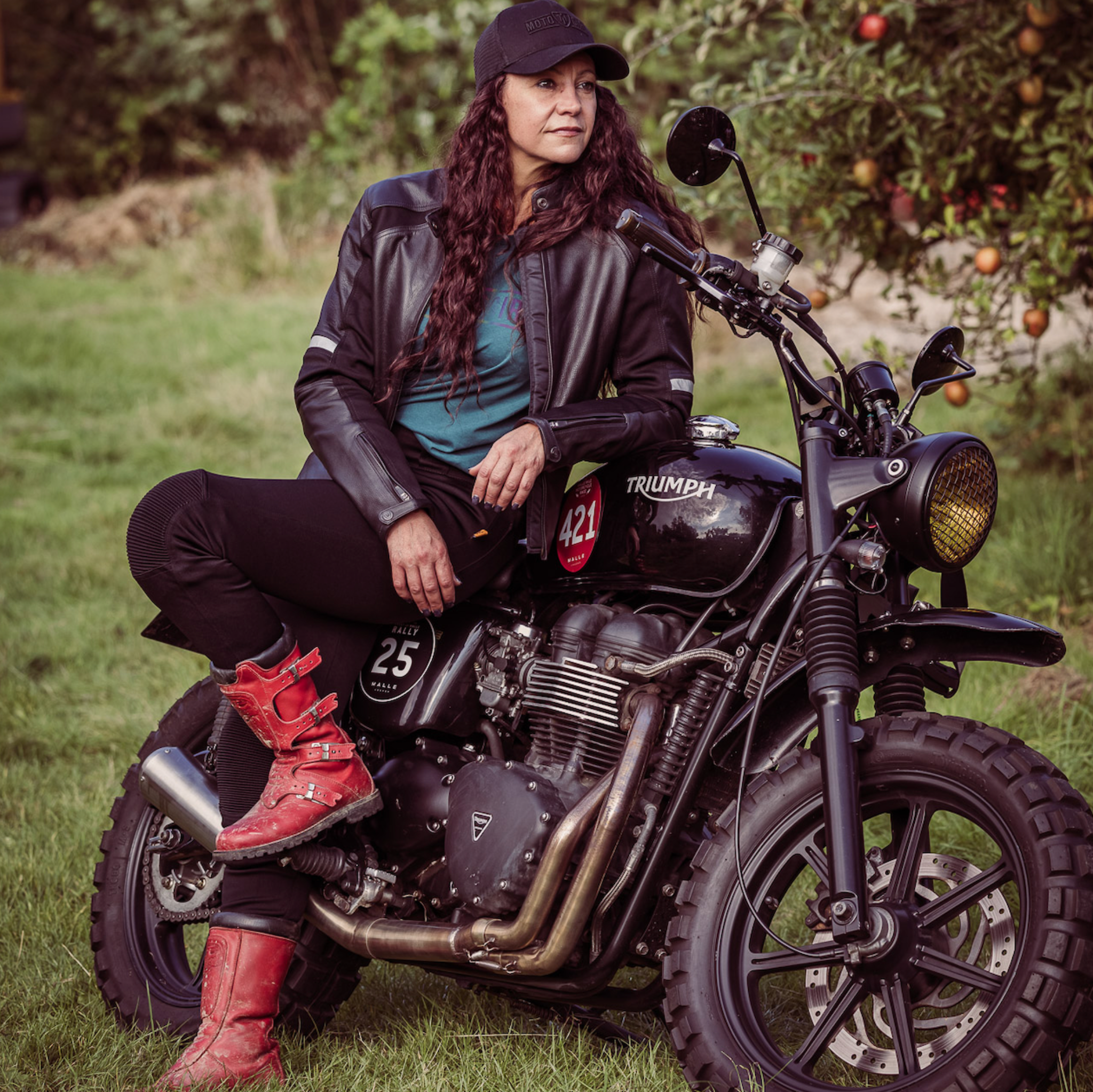 A woman leaning on her Triumph motorcycle wearing black leather motorcycle jacket and red boots 