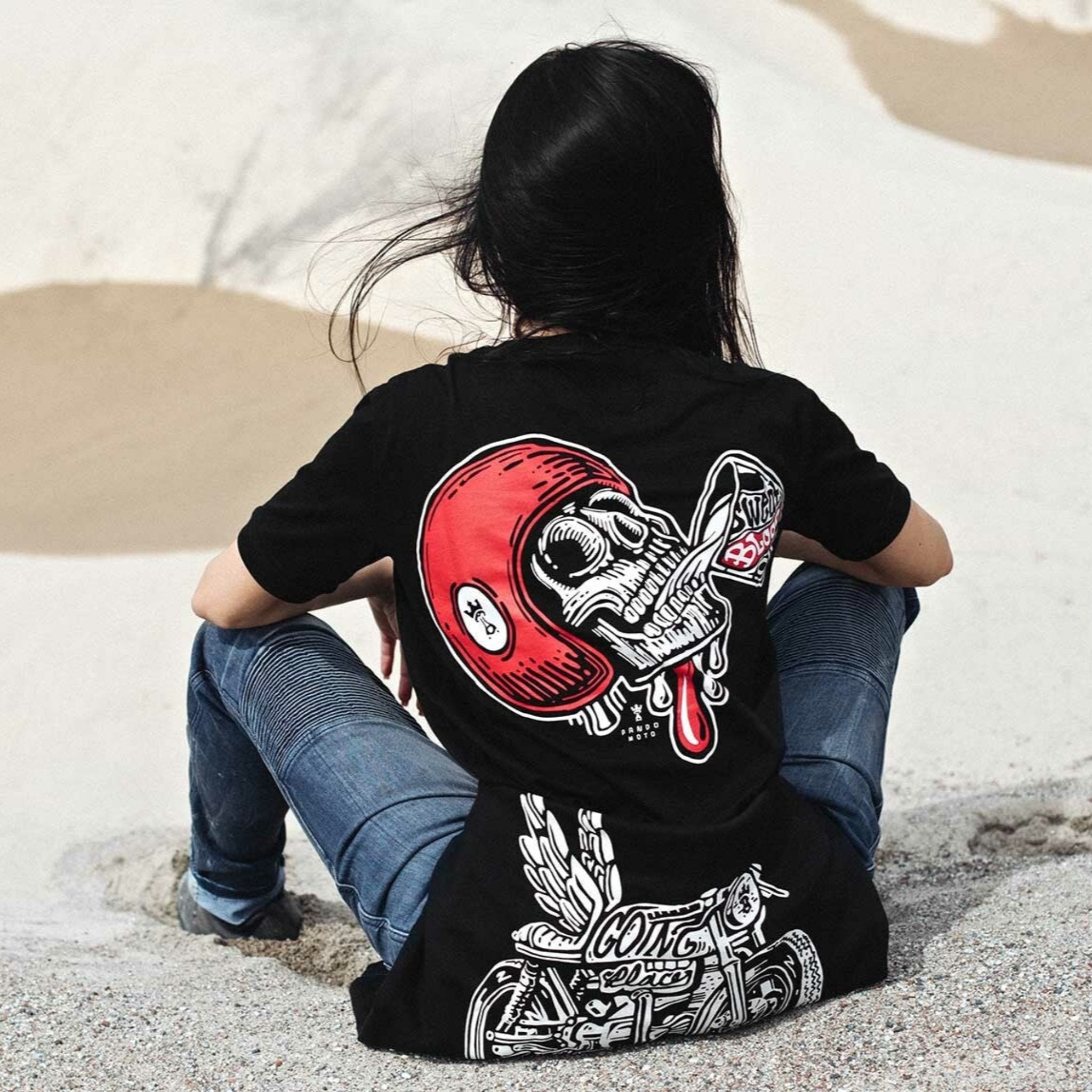 A woman siting on the beach wearing black Pando Moto women's motorcycle t-shirt with red scull motive