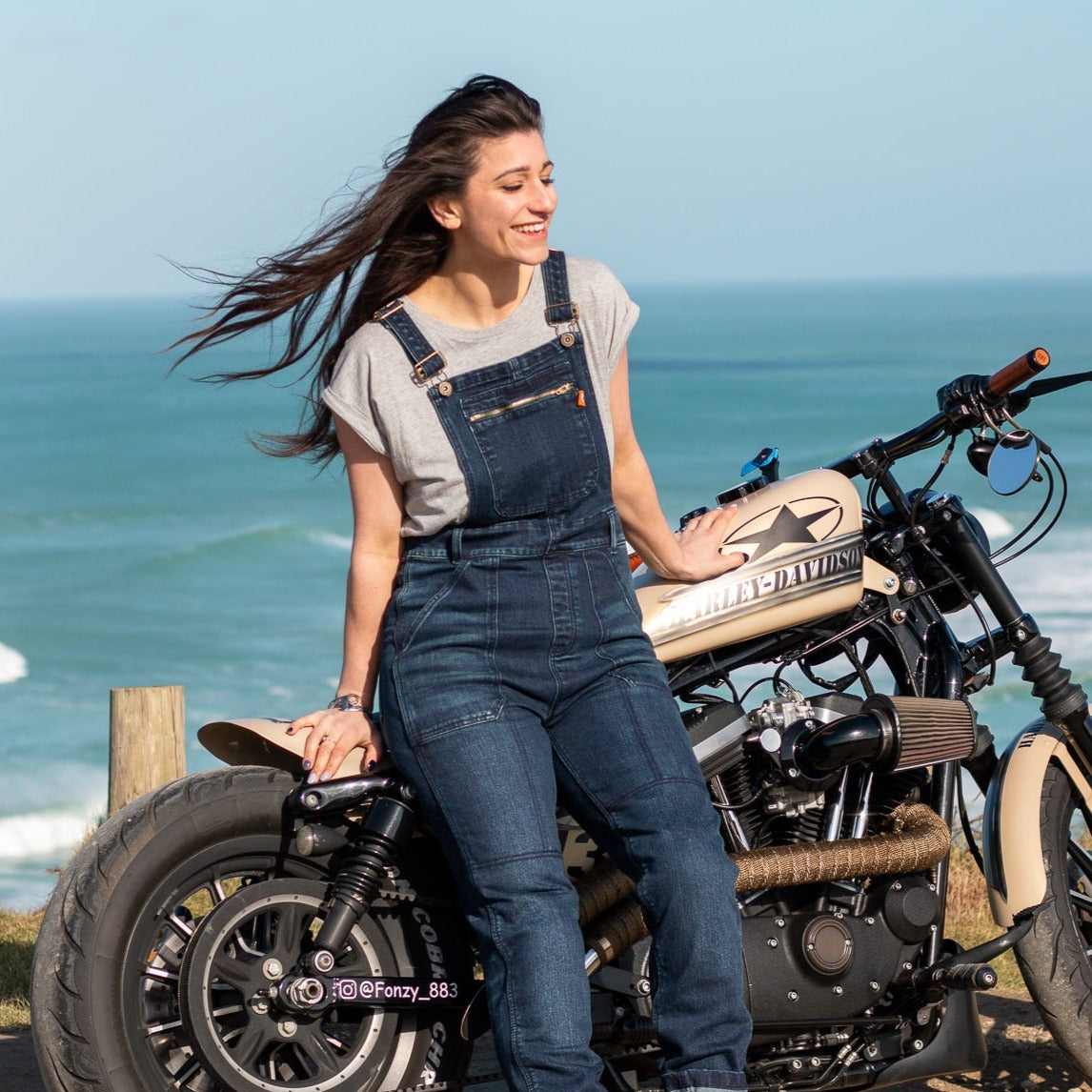 A woman by the beach with her motorcycle wearing blue women&#39;s motorcycle overall from Moto Girl