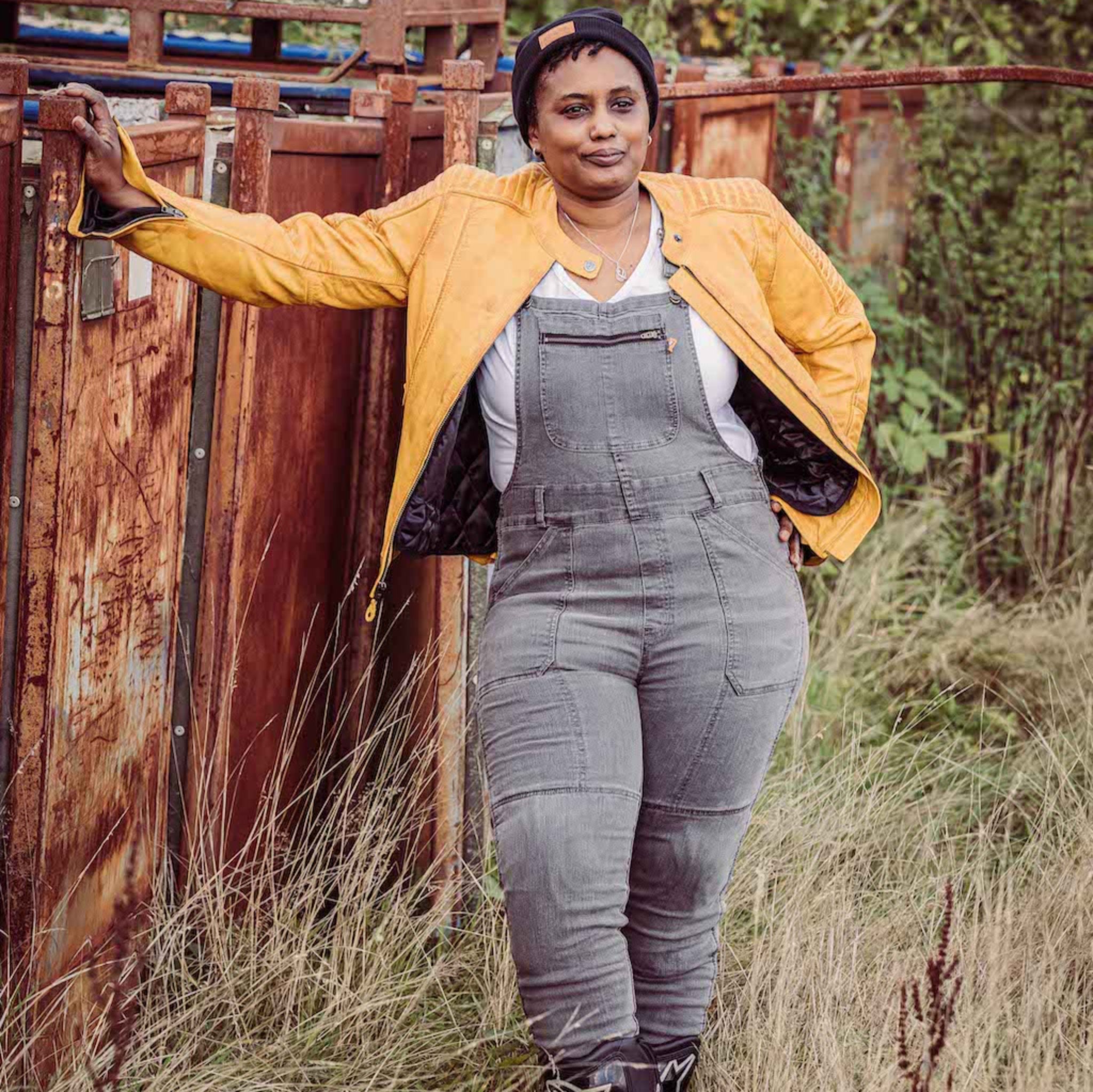 A woman wearing grey motorcycle overall and yellow leather jacket standing by the metal container