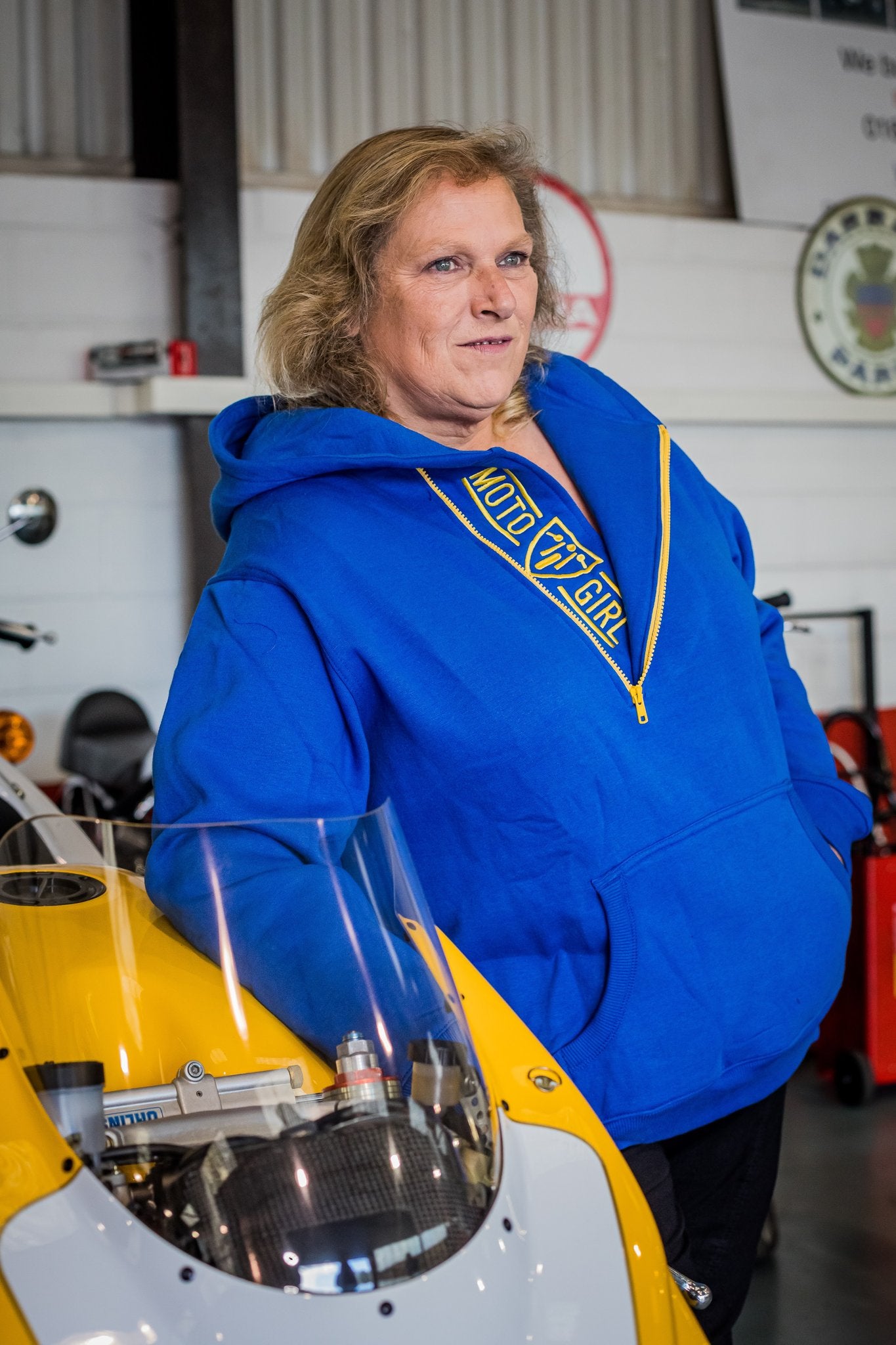 A blond woman standing by a motorcycle wearing blue and yellow motorcycle hoodie from Moto Girl