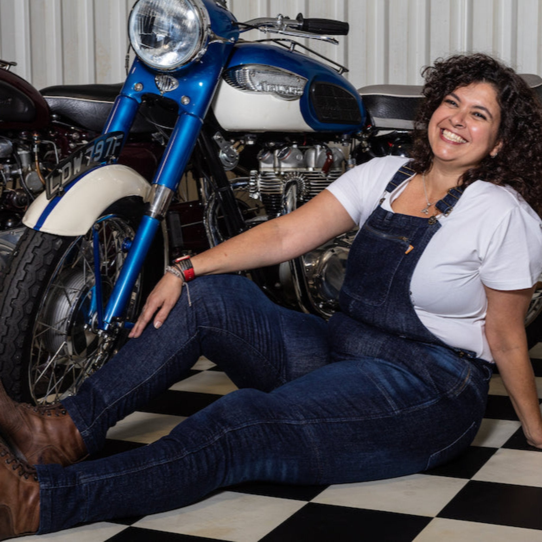 A smiling woman siting on the floor in the motorcycle shop and wearing blue motorcycle overall 