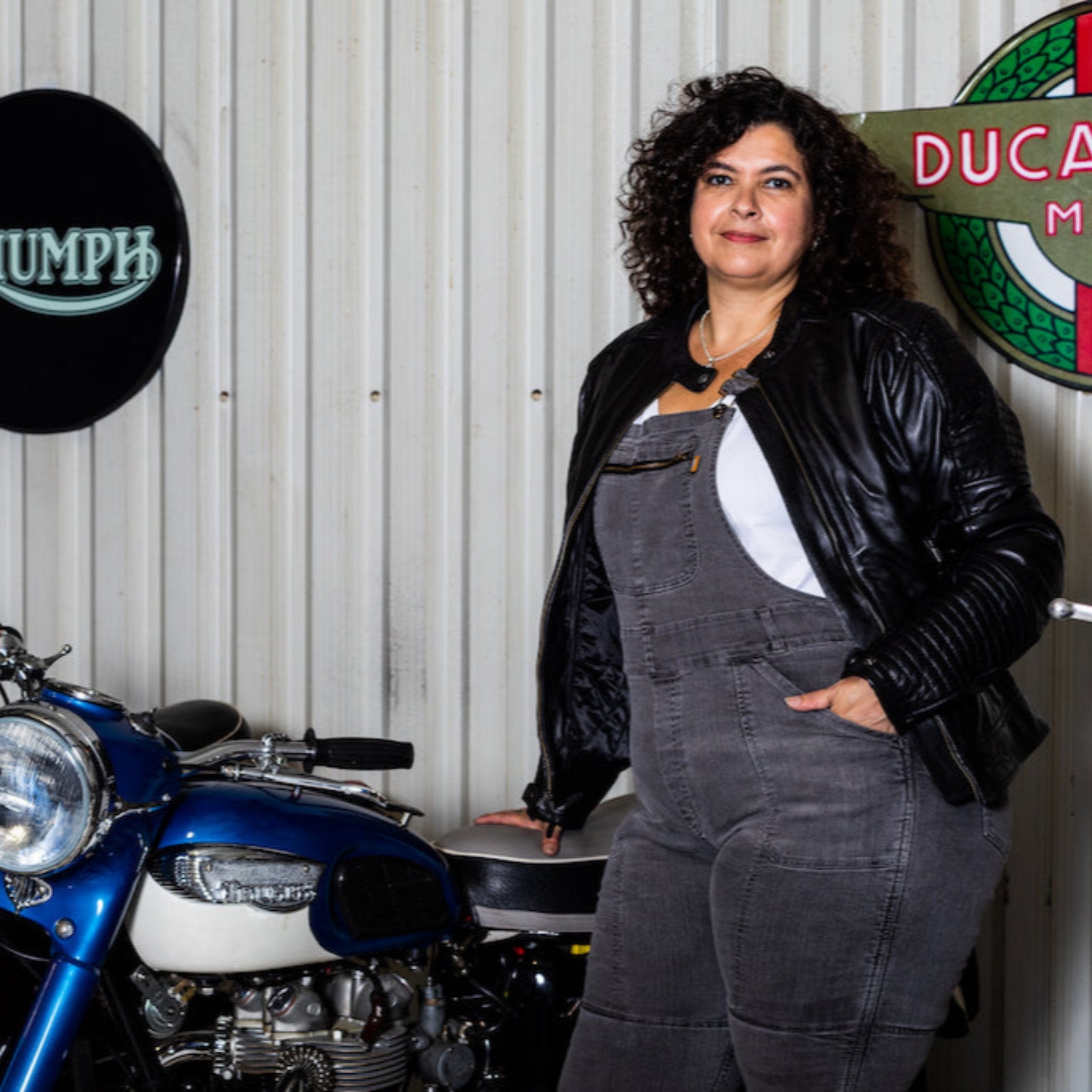 A woman wearing black motorcycle leather jacket and grey overall standing by a motorcycle