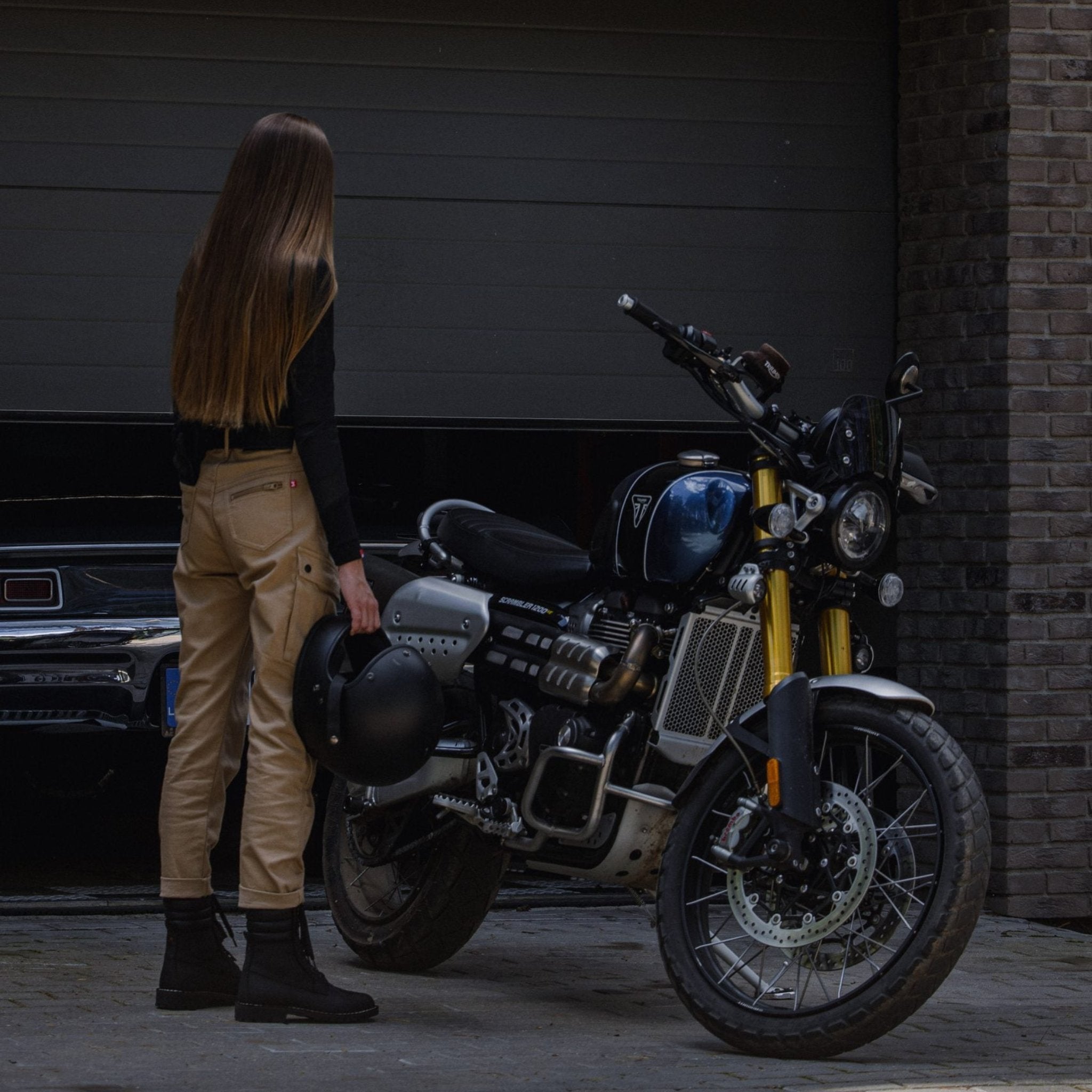 A brunette woman wearing beige Cargo Mila pants, standing next to her motorcycle and holding her helmet, viewed from the back.