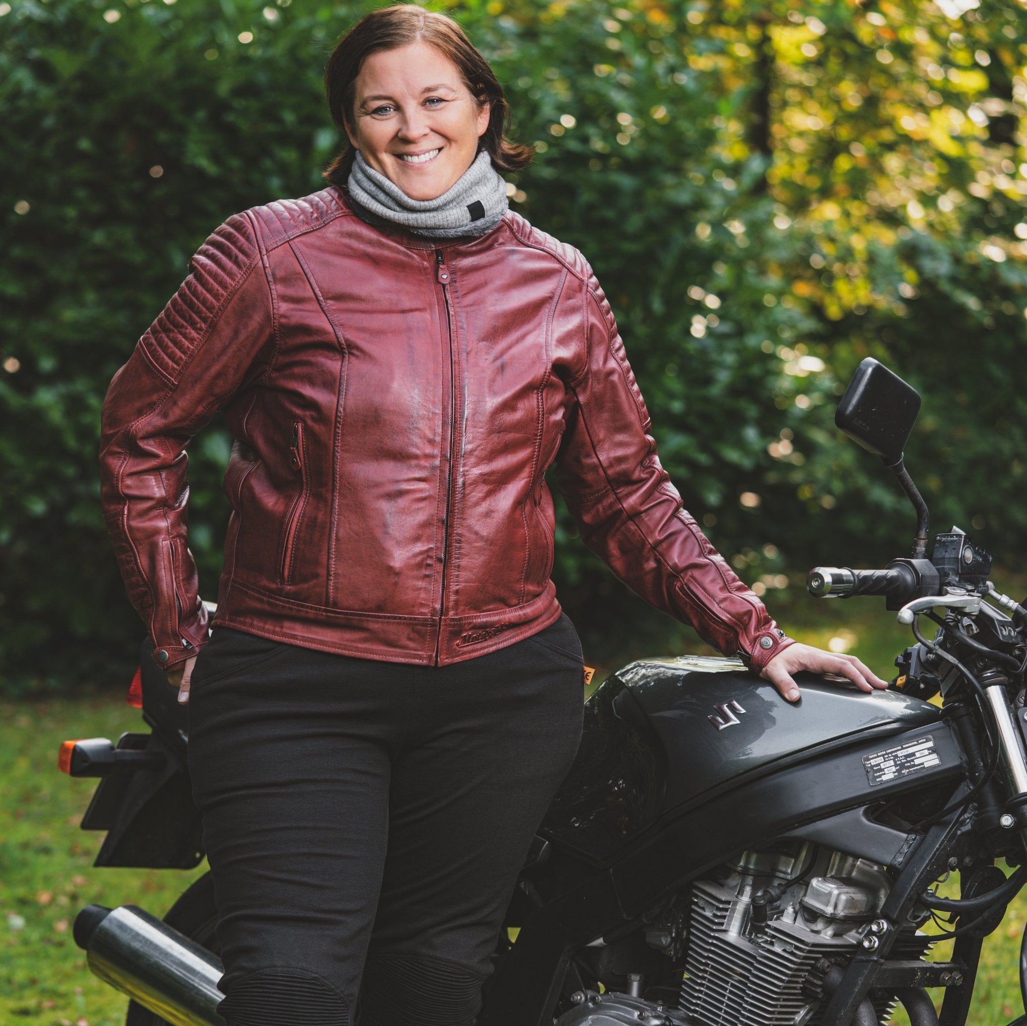 smiling woman standing next to a Suzuki motorbike wearing red Valerie mc jacket from moto girl 