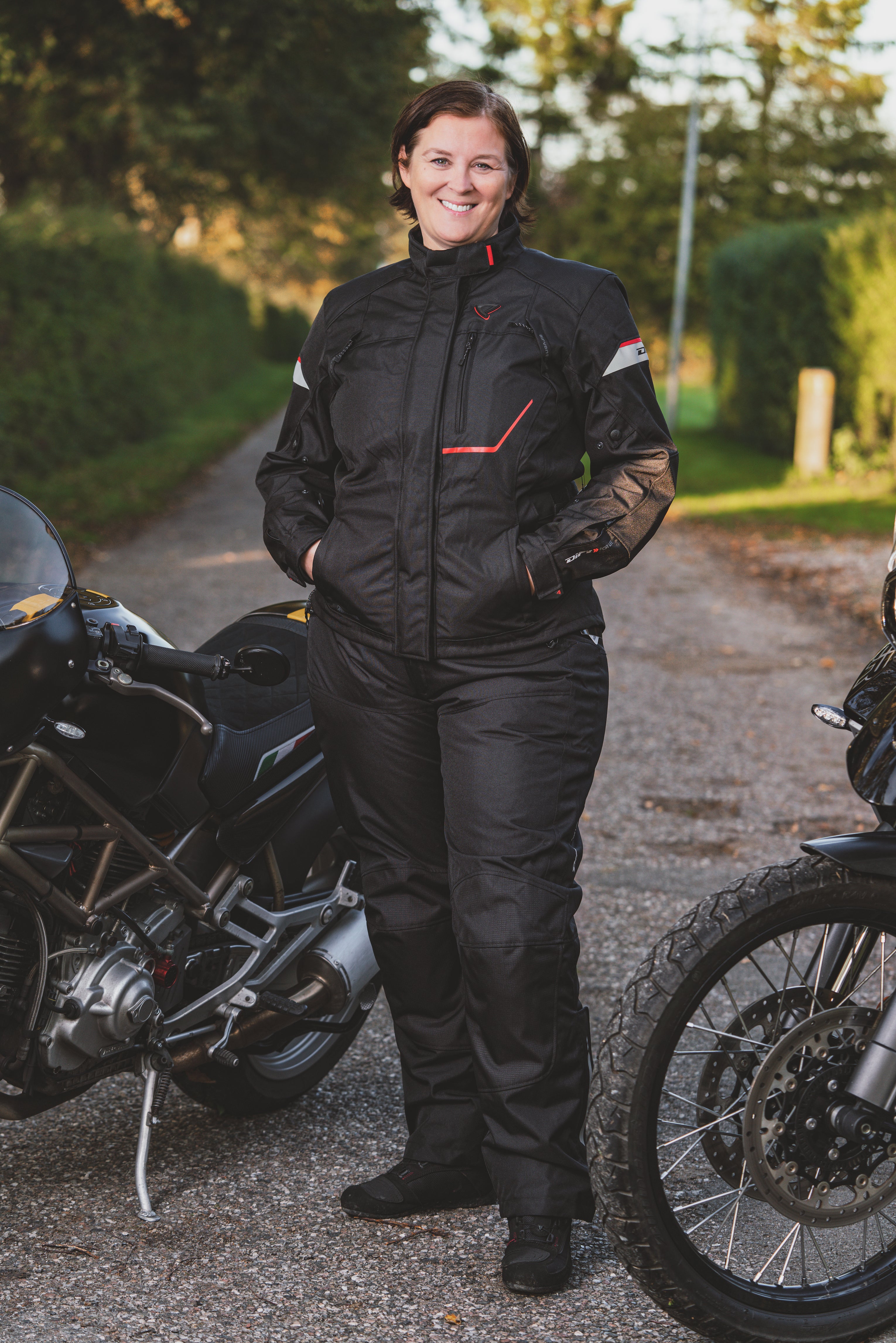A smiling woman standing by motorcycles wearing DIFI motorcycle black Cyclone pants and Nexia jacket
